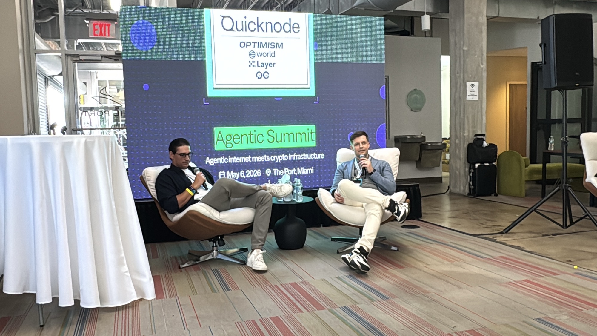 Fireside chat at the Agentic Tooling Summit at The Port, Miami, with the QuickNode / Optimism / World / X Layer / 0G sponsor band on the LED screen behind the stage