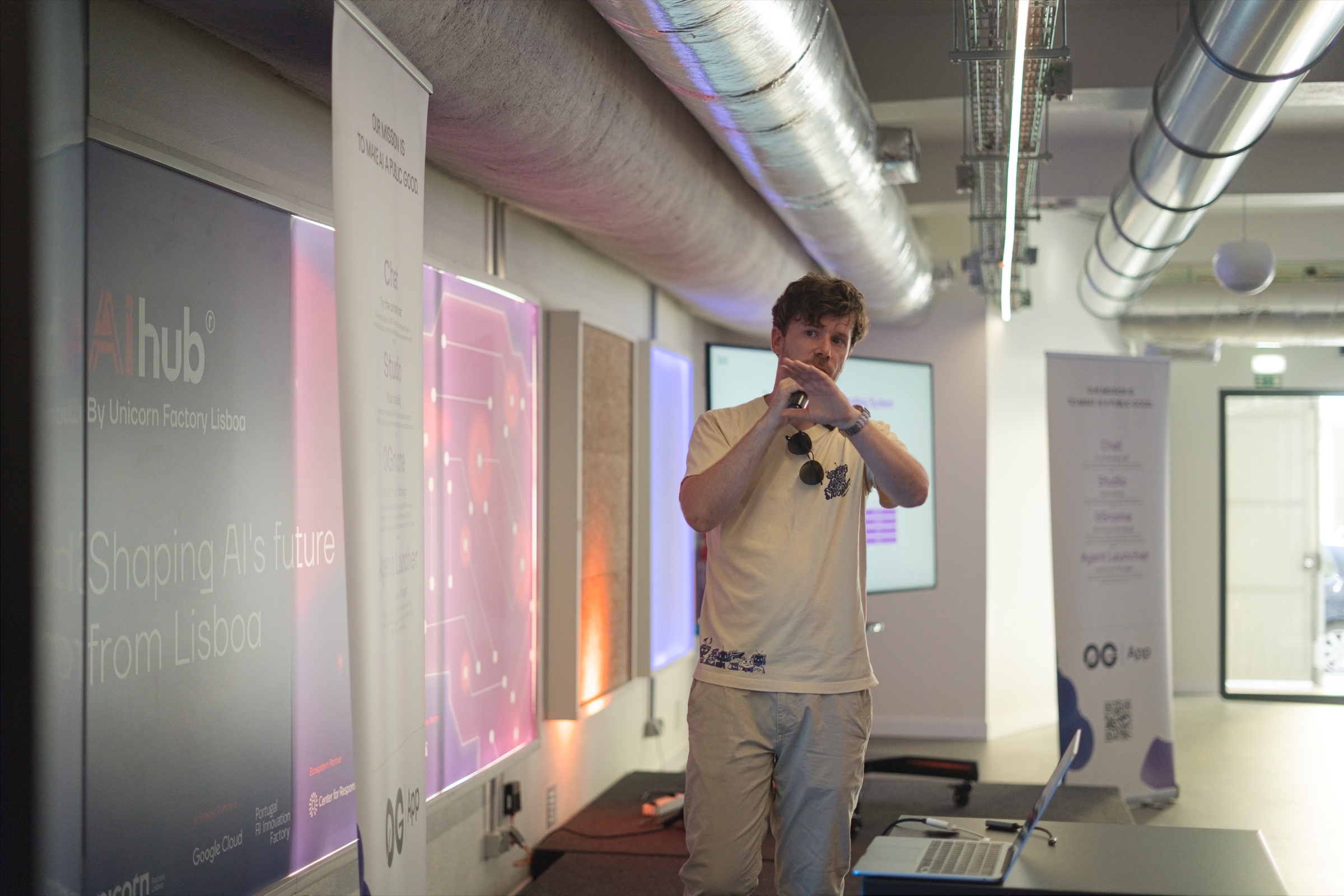 Speaker with a microphone presenting between AI Hub and 0G App banners at the House of AI Demo Day in Lisbon